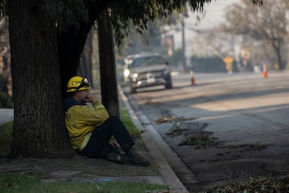 Un bombero descansa en el suelo mientras varios de sus compañeros refrescan un edificio humeante en una calle de Pacific Palisades, en Los Ángeles.